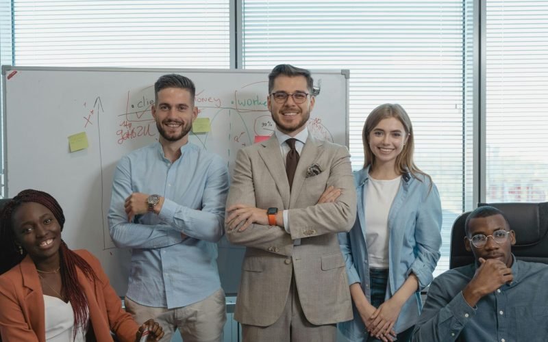 Five coworkers smiling and posing in a bright modern office with a whiteboard and window view.